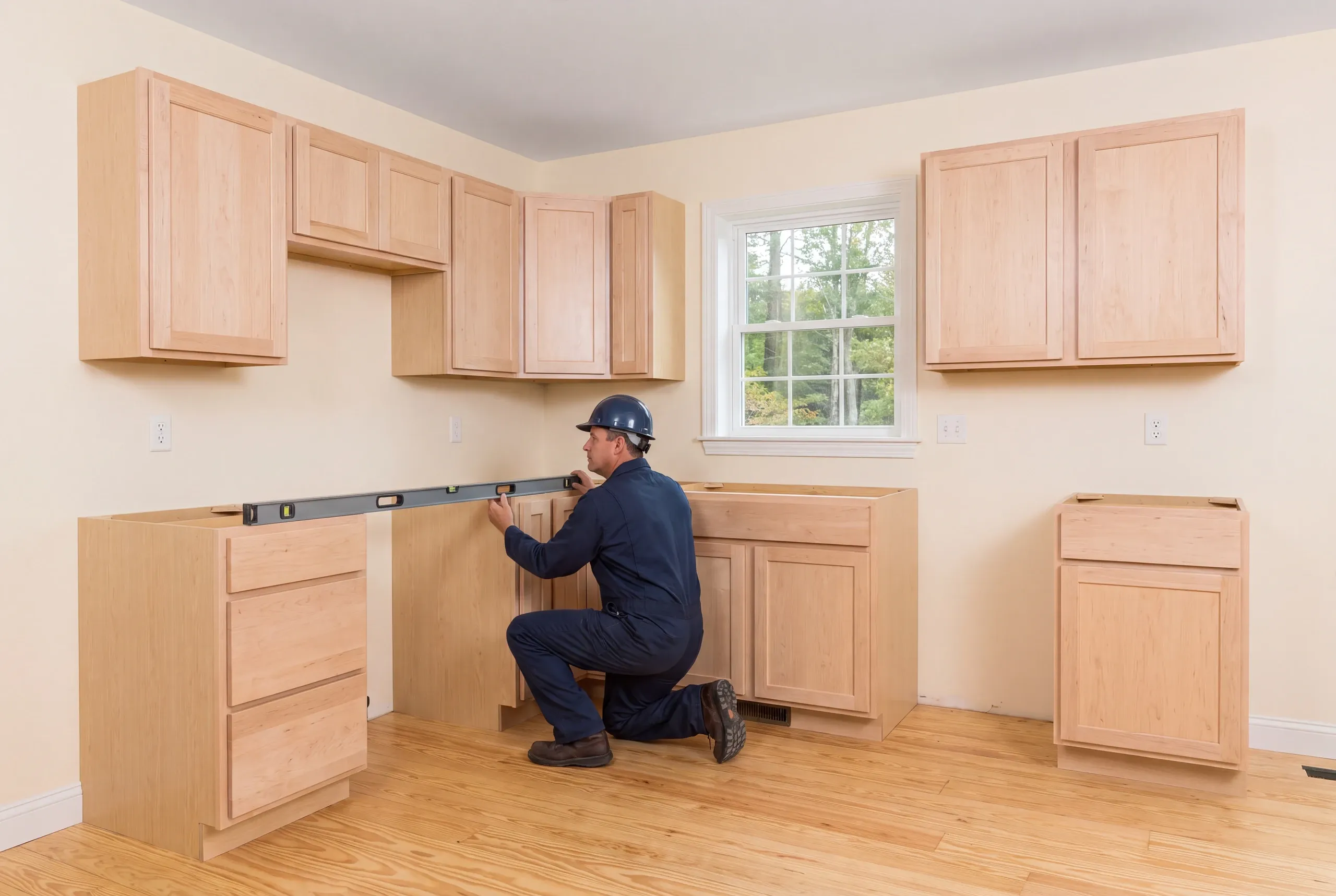 A worker in a hard hat kneels on a wooden floor in a kitchen, holding a long level across base cabinets.