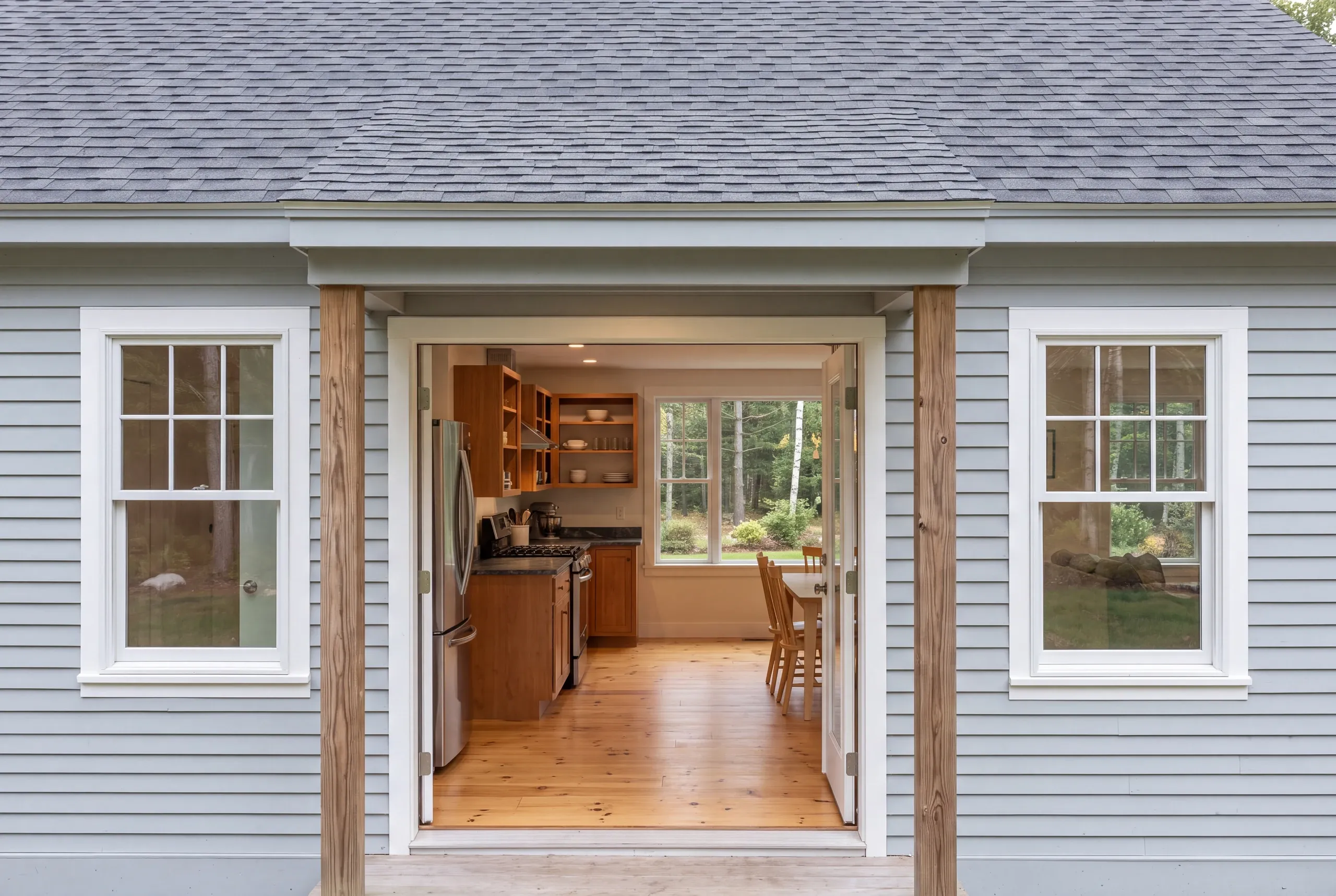 Exterior view showing light blue siding, gray shingled roof, two multi-pane windows, and an open doorway looking into a kitchen.