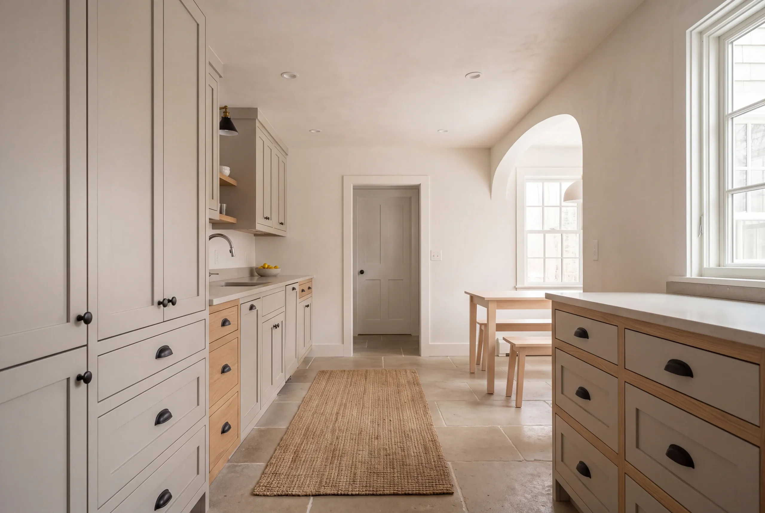 A kitchen featuring light shaker cabinetry, a central walking aisle with a runner rug, and a dining table set in an adjoining arched alcove.
