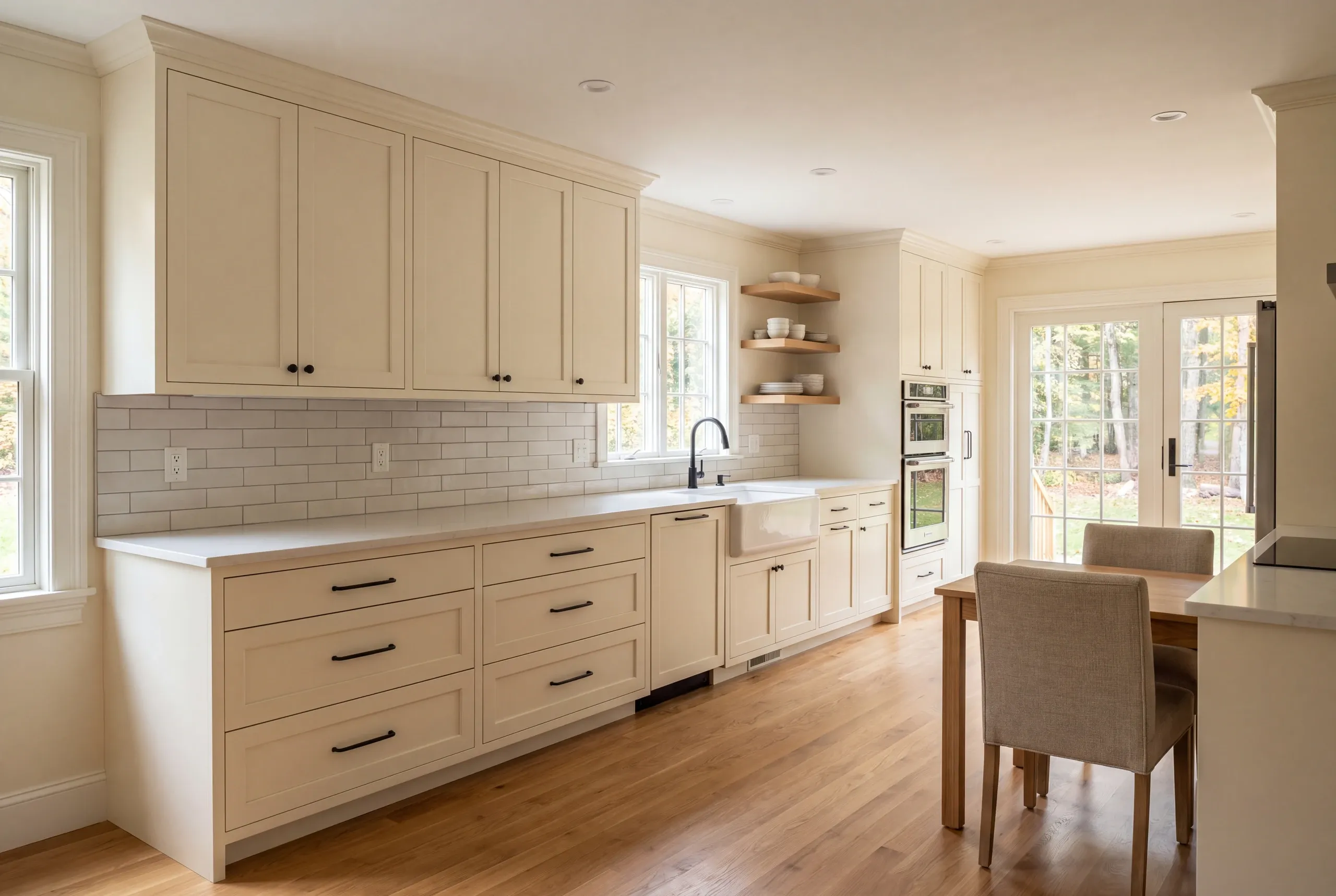A bright kitchen interior features light-colored shaker cabinets, a pale countertop, white tile backsplash, and a dining set in the corner.