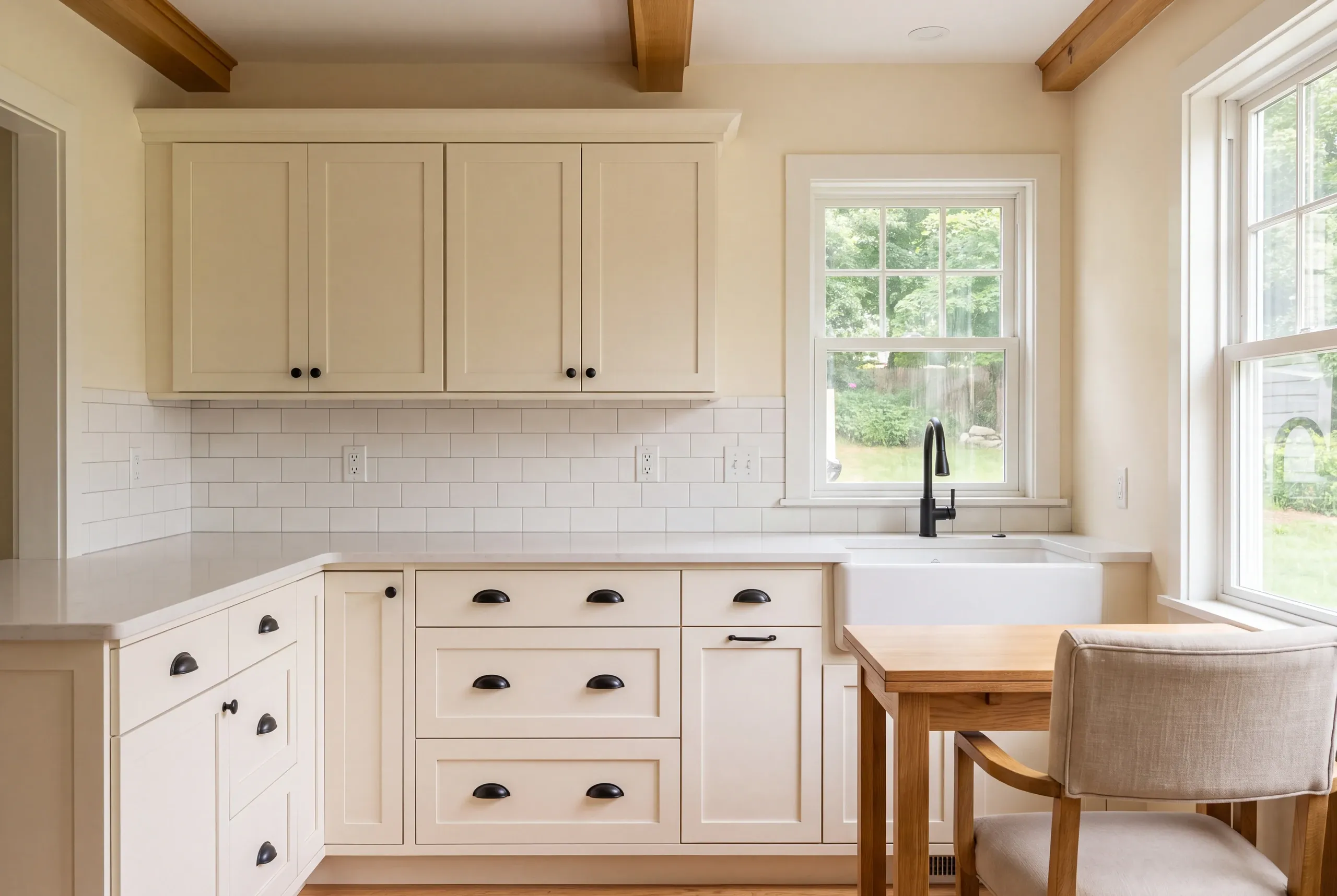 Cream-colored kitchen cabinets with dark hardware, a white tile backsplash, and a dining table with a chair are visible.