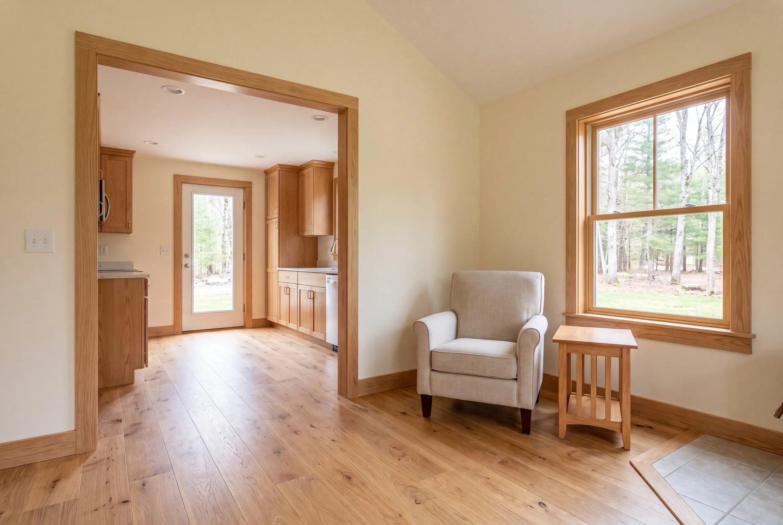 An armchair and small side table sit on a light wood floor in a brightly lit room connected by a wide oak-trimmed doorway to a kitchen.